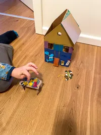 A house made of paperboard and painted colorful with watercolors. The is house standing on the floor.