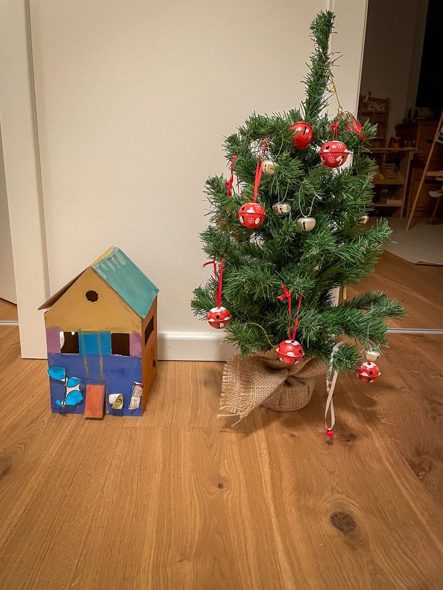 The paperboard house is standing next to small christmas tree that is double as tall than the house.