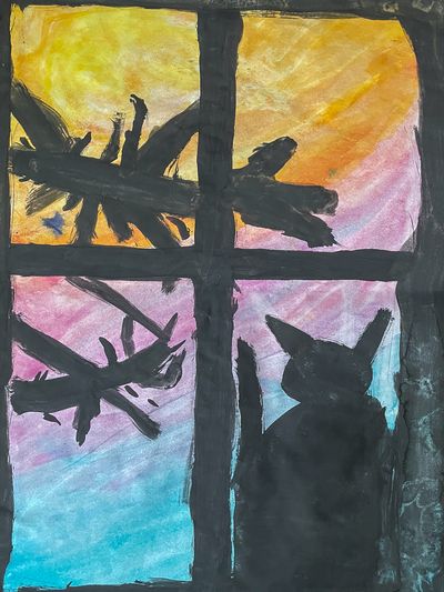 A black cat sitting in front of a window watching the yellow-orange-cyan sky and black tree branches.