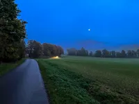 An intense dark blue evening sky with a small street and a car with lights on surrounded by green fields.