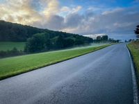 A winding, uphill road with a green, misty pasture on the left side