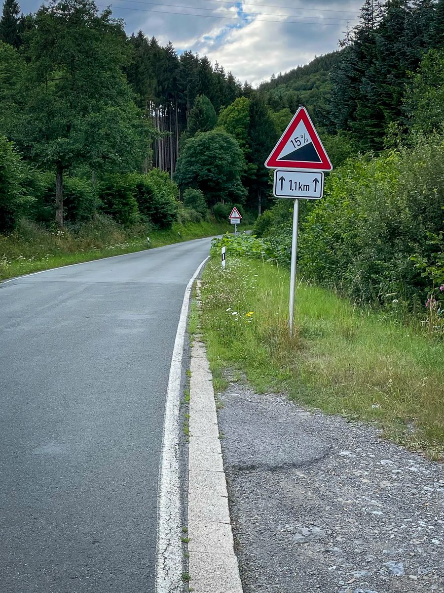 A street sign saying '15% incline' and a small road pointing up into a dark forest