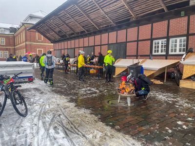 An outdoor backyard of the Nordkirchen castle in a wet and snowy setting cyclists are gathering around a campfire and catering desks.