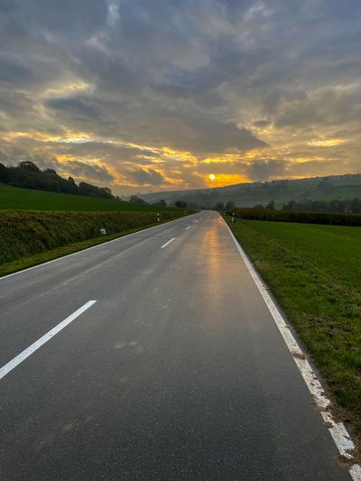 A wet road surrounded by green hills pointing into the evening sunset with dramatic skies.