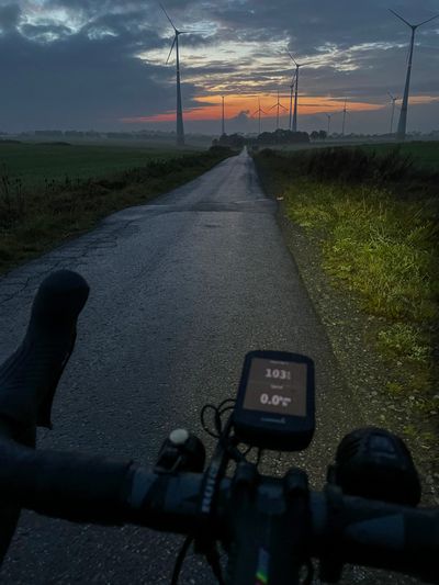 A picture with the drop handlebars pointing down a small and dark road, surrounded by wind turbines. The road leads into the final of an orange sunset with intense clouds.